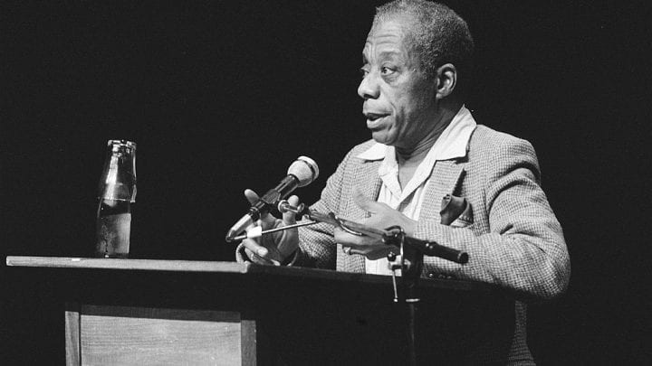 James Baldwin speaking from a lectern with microphone, black and white